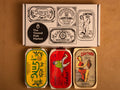 Three colourfully packaged tins of fish beneath a gift box illustrated with line drawings of the tins and The Tinned Fish Market logo. Against a light brown background.