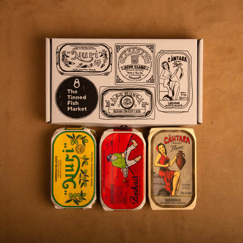 Three brightly-coloured tins of Portuguese tinned fish below an illustrated cardboard gift box, against a light brown background.