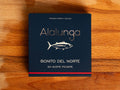 Canned white tuna in square blue card packaging with Alalunga in silver, embossed lettering above a diagram of a tuna. There is a red stripe along the bottom of the packaging.