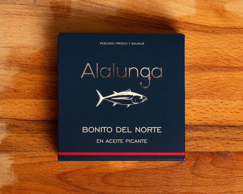 Canned white tuna in square blue card packaging with Alalunga in silver, embossed lettering above a diagram of a tuna. There is a red stripe along the bottom of the packaging.