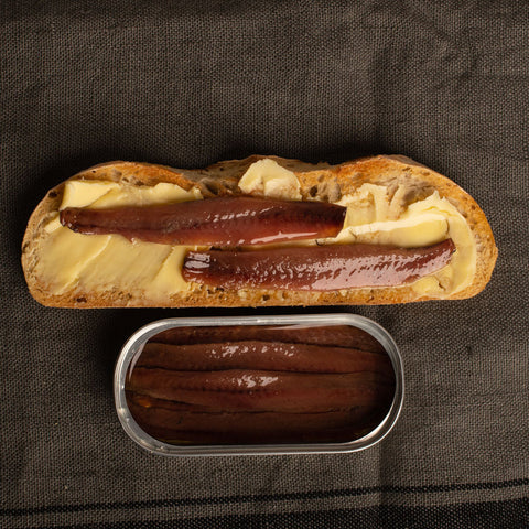 An open tin of deep-brown anchovies beneath a slice of buttered toast with two anchovy fillets, on a grey tea-towel backdrop.
