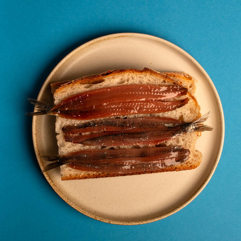 Some anchovies gently placed on top of toast on a golden trim plate with an aqua blue background.
