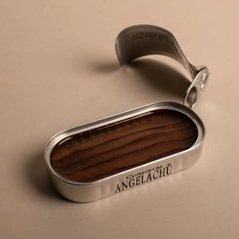 An open tin of anchovies with brown anchovy fillets in olive oil inside and conservas Angelachu written on the side of the silver-coloured tin. The lid with ring-pull attached is by its side.