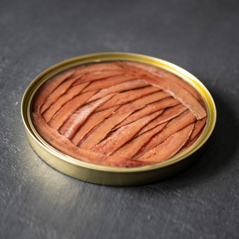 An open circular gold tin of anchovy fillets against a grey background