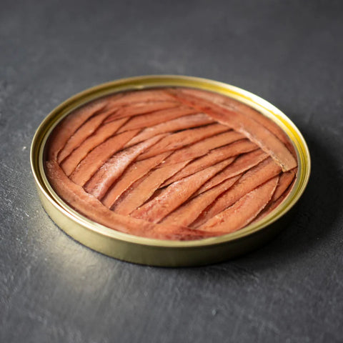 An open circular gold tin of anchovy fillets against a grey background.