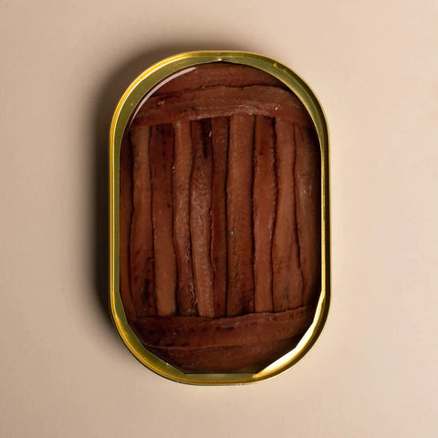 An open tin of anchovy fillets. The fillets are a deep brown and are framed at either end by shorter fillets, against a light background.
