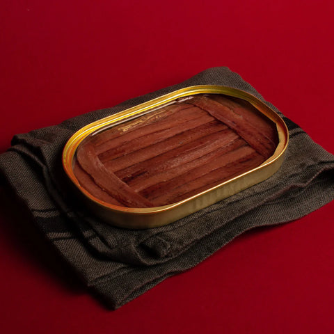 An open tin of anchovy fillets. The fillets are a deep brown and are framed at either end by shorter fillets, against a vibrant red backdrop.