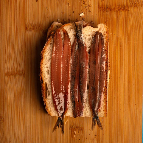 Three tasty-looking butterflied anchovy fillets resting on a fresh slice of bread. A light brown wooden table as the backdrop.
