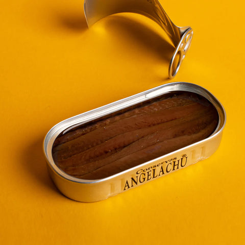 A silver-coloured tin of deep brown anchovies, the peeled off lid by its side, against a yellow backdrop.