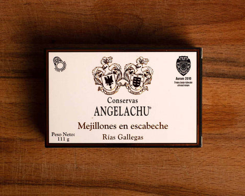 A tin of mussels in rectangular white card packaging with a black border with Angelachu written below two coats of arms