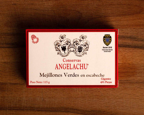 A tin of mussels in rectangular cardboard packaging. The packaging is white with an orange border. There are two coats of arms above Conservas Angelachu, which is written in orange lettering.