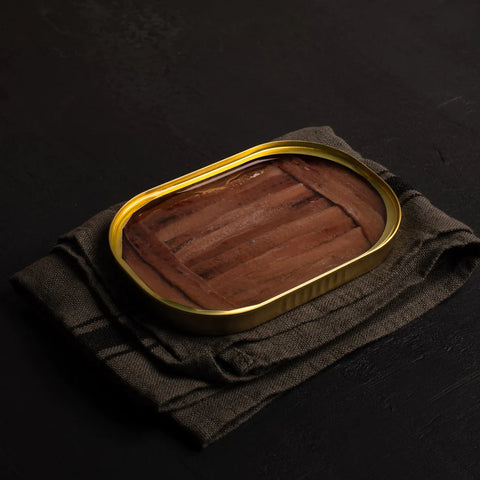 An open gold-coloured tin of anchovy fillets. The fillets are a deep brown and are framed at either end by smaller fillets. Neatly resting on a folded grey-black cloth on top of a black countertop. An open gold tin of anchovy fillets. The fillets are a deep brown and are framed at either end by smaller fillets. neatly resting on a folded dark grey cloth on top of a black countertop. 