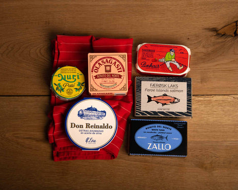 Six brightly coloured tins of fish, three of them on a red tea towel, against a wood background