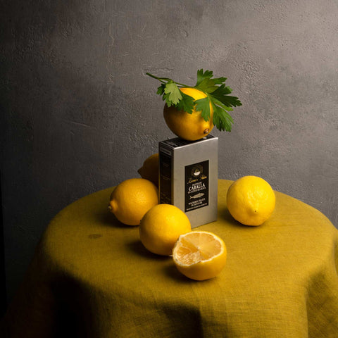 A tin of mackerel in silver packaging with lemons around it and one lemon balanced on top of the tin on a yellow tablecloth against a grey background 
