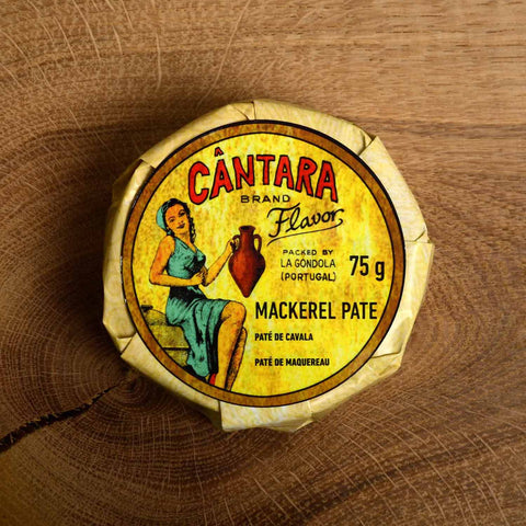 A round tin of mackerel pâté in a glossy paper wrap with an image of a woman in a green dress holding an amphora of olive oil on the front