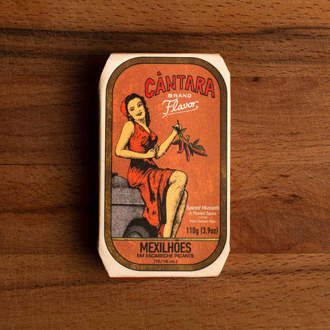 A tin of spicy mussels in an orange paper wrap with a woman in an orange dress holding a bunch of chilli peppers beneath the Cântara logo written in red.