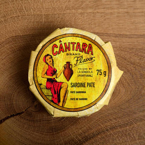 A round tin of sardine pâté in a glossy paper wrap with an illustration of a woman in a red dress beneath Cântara written in red lettering
