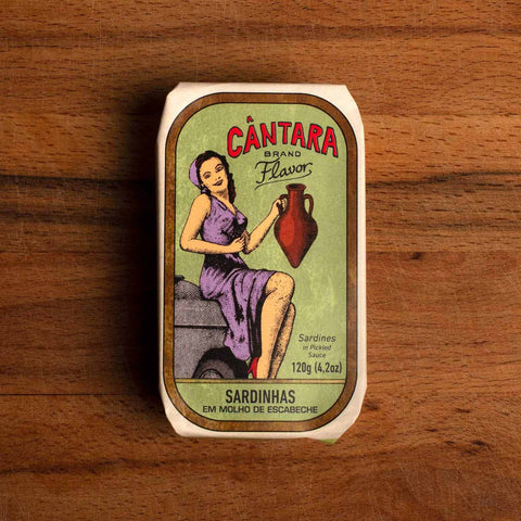 A tin of sardines in a paper wrap depicting a woman in a purple dress holding an amphora of olive oil. The tin is on a wood background.