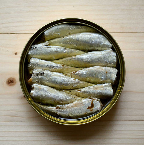 An open tin of carefully arranged bright silver small sardines. The tin is on a pale pine background.