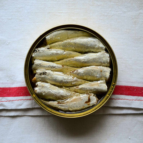 A round tin of carefully arranged small sardines in golden olive oil. The tin is resting on a linen tablecloth.