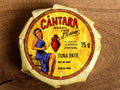 A round tin in a glossy paper wrap with a woman on the front in a blue dress holding a container of olive oil beneath the Cântara logo in red.