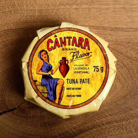 A round tin in a glossy paper wrap with a woman on the front in a blue dress holding a container of olive oil beneath the Cântara logo in red.