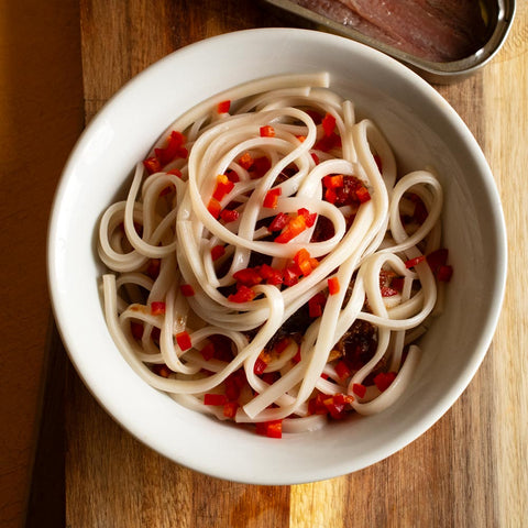 A bowl of spicy noodles topped with red peppers and accompanied by an open tin of anchovy fillets.