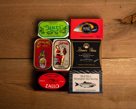 Seven tins of brightly packaged tinned fish against a wood background