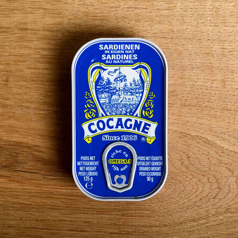 A blue tin with Cocagne written on the front beneath an illustartion of the game tallow stick. The tin is against a wood backdrop.