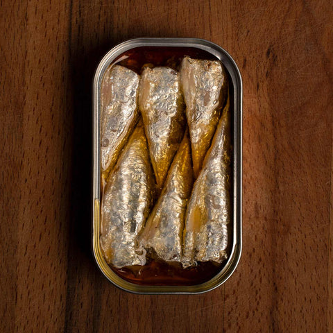 Six small sardines in chilli, carefully arranged in a tin, on a dark brown table. Waiting to be enjoyed.