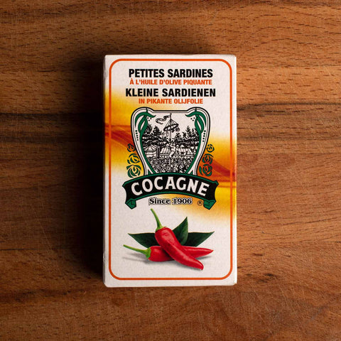 A tin of spicy sardines in a rectangular box. There are two red chilli peppers. Above the image Cocagne is written in white on a black and green banner. There is a drawing of the tallow-stick game above the logo.