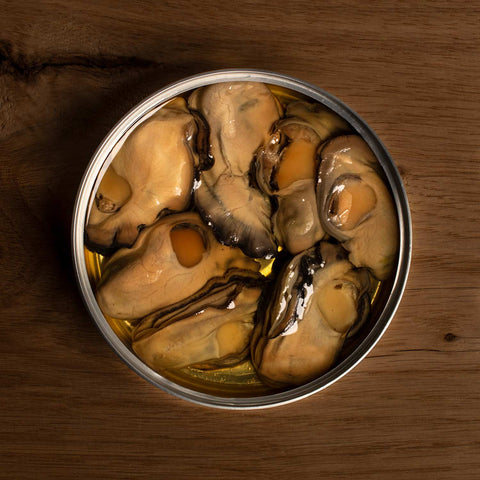 An open, round tin of seven large oysters in olive oil, on a wood background.
