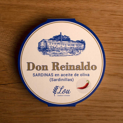 A round tin with a white card wrap. The cover has a blue and gold border. Don Reinaldo is written in gold lettering below a drawing of the cannery. and above a red chilli pepper.