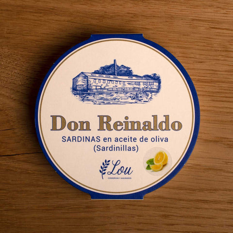 A round tin of sardines with a white card wrap. There is an image of a lemon in the corner, above which Don Reinaldo is written in gold lettering beneath a drawing of the cannery.