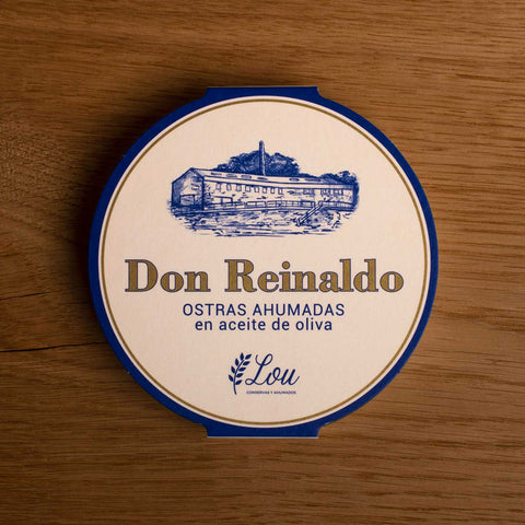 A round tin with a white card wrap. The cover has a blue and gold border. Don Reinaldo is written in gold lettering below a drawing of the cannery.