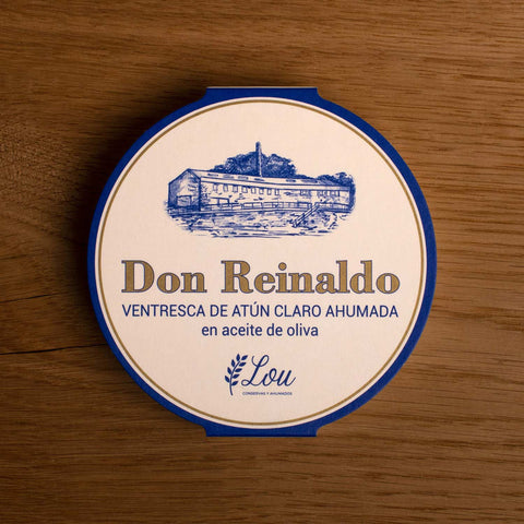 A round tin with a white card wrap. The cover has a blue and gold border. Don Reinaldo is written in gold lettering below a drawing of the cannery.