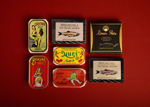 This box is a simple luxe edit of sardines and smoked sprats. The tins include unique illustrations giving them character, such as a fisherman sitting barefoot mending a fishing net, an elegant lady, and images of sardines.