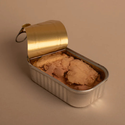 An open tin of cod liver. The tin is silver. The peeled back lid is gold. On a beige-coloured background.