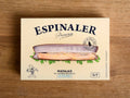 A tin of razor clams in a rectangular white box. On the front, there is an image of a razor clam resting on some green parsley leaves with Espinaler written in black lettering above.