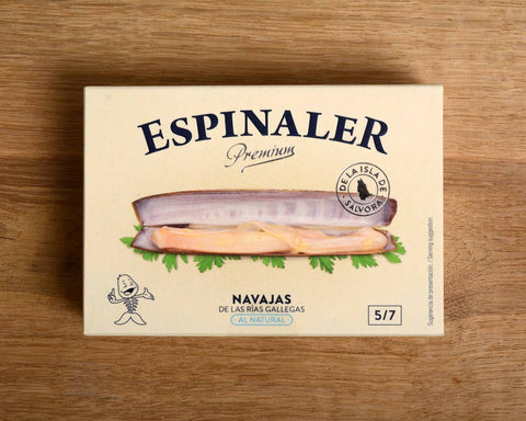 A tin of razor clams in a rectangular white box. On the front, there is an image of a razor clam resting on some green parsley leaves with Espinaler written in black lettering above.