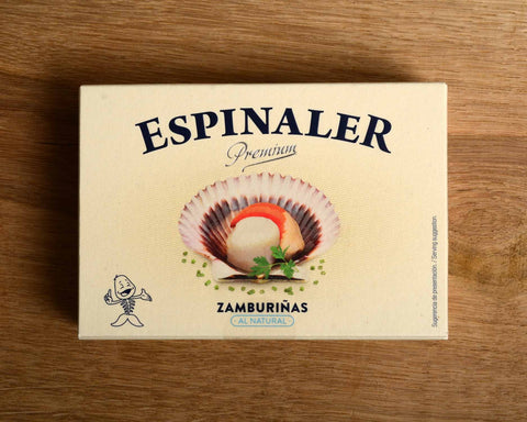 A cream-coloured rectangular box with an open variegated scallop in its shell on the front, beneath Espinaler written in black lettering. The tin is on a wood background.