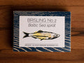 A tin of sprats in a rectangular box with a blue border and a painting of a yellow sprat on the front, against a dark wood background.
