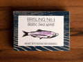 A tin of sprats in a rectangular box with a blue border and a painting of a purple sprat on the front, against a dark wood background.