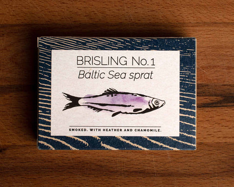 A tin of sprats in a rectangular box with a blue border and a painting of a purple sprat on the front, against a dark wood background.