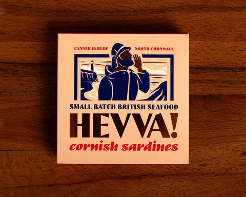 Box containing Cornish sardines from Hevva! The box is illustrated with a fisherman calling out over the Cornish coast with cliffs and a lighthouse in the background. The box is colourful with Hevva! written in golden, embossed lettering  
