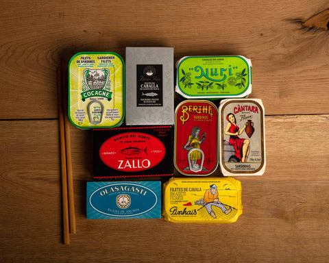 Eight tins of fish in brightly coloured packaging with a pair of chopsticks next to them on a wood background