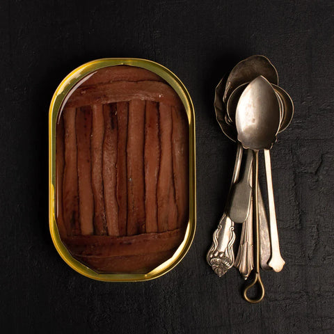 An open tin of anchovy fillets. The fillets are a deep brown and are framed at either end by shorter fillets. Gold trim around the tin with a number of short silver spoons by its side set off by a striking black backdrop.