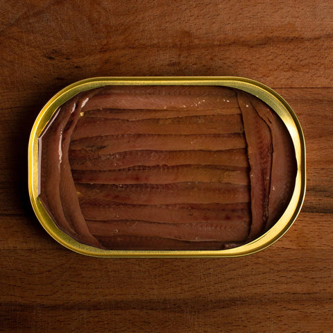 An open gold-coloured tin of anchovy fillets. The fillets are a deep brown and are farmed at either end by shorter fillets, against a dark wooden table.