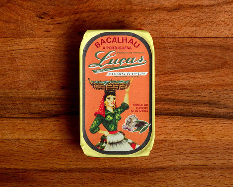 A tin of salt cod with orange packaging, a yellow border and an image of a woman dressed in green, red and white carrying a basket of fish balanced on her head 