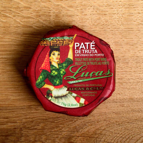 A round tin in a port wine-coloured paper wrap with Luças written in green lettering next to a woman in red, green and white carrying a basket of fish